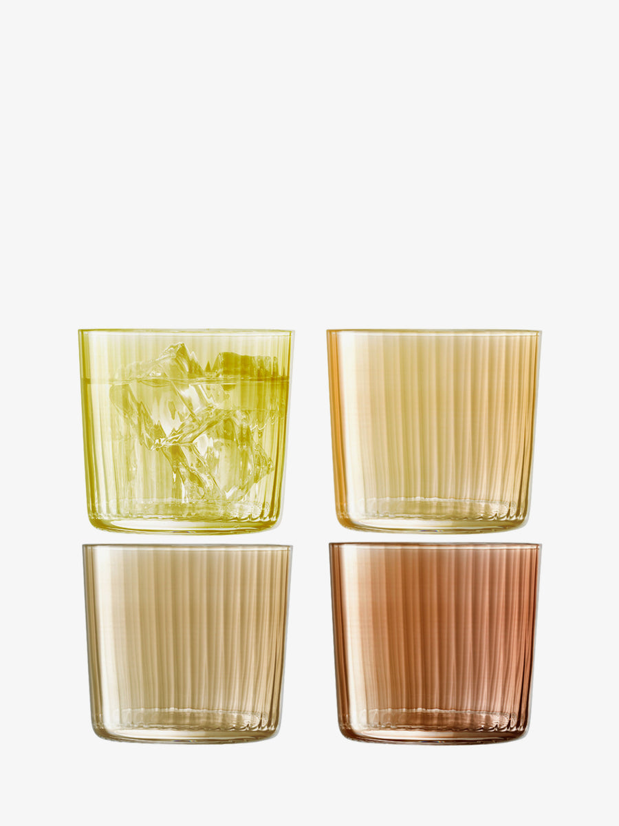 Four ribbed fluted Tumbler glasses by lsa-international in yellow, gold, rose, and amber. The yellow glass is filled with ice while the others remain empty. Displayed on a plain white background, this vibrant set of stackable tumblers from lsa-international is perfect for any occasion.