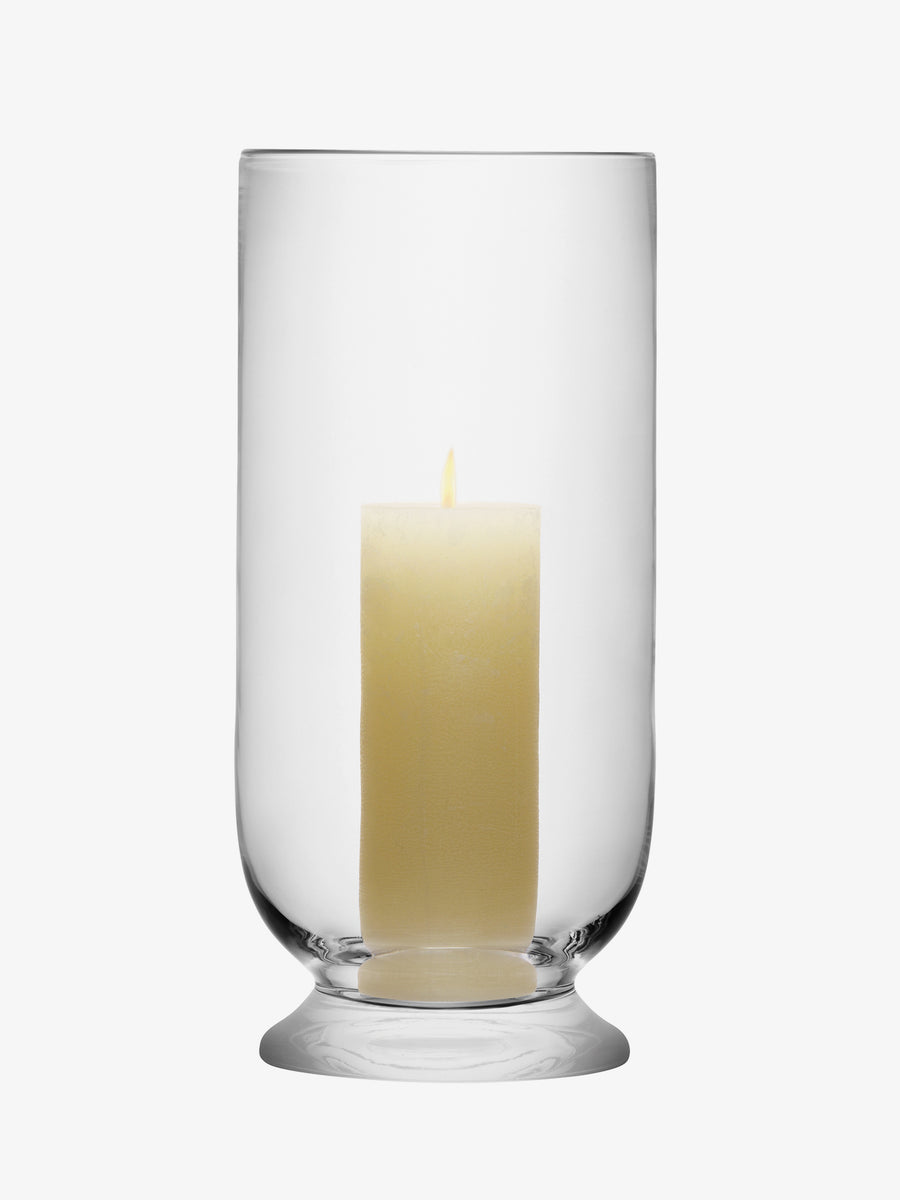 A lit cream-colored pillar candle sits inside the clear, cylindrical Storm Lantern by lsa-international against a plain white background.