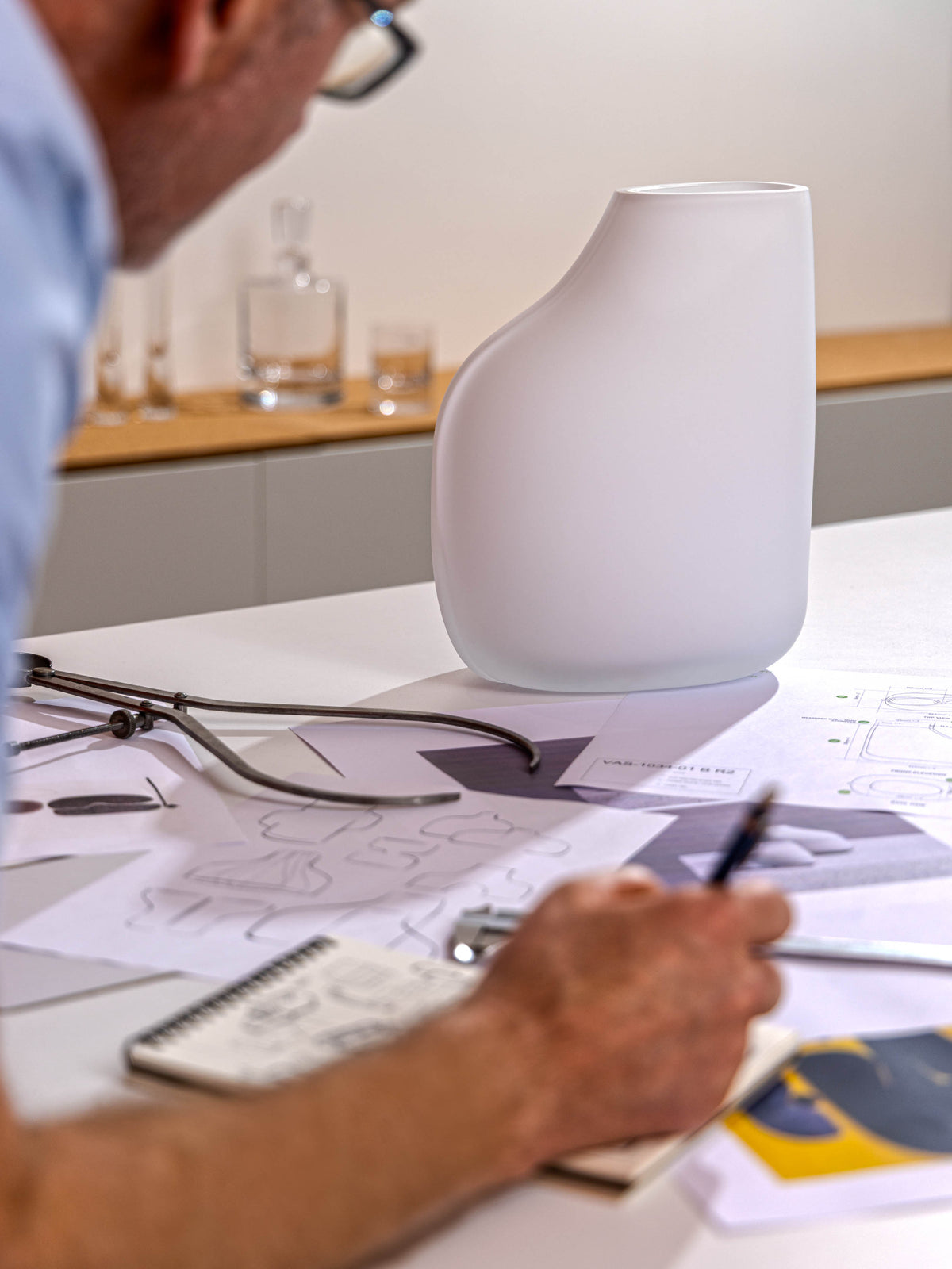A person sketches design ideas in a notebook on a desk covered with blueprints and tools, with the 1966 Vase by lsa-international—a modern, white, abstract-shaped limited edition vessel and handmade collector’s piece—in the background.