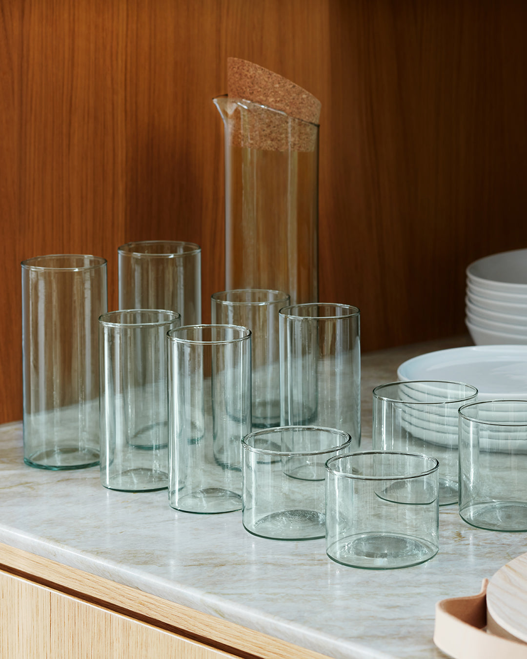 Nine lsa-international Tumbler glasses, featuring a short cylindrical design and crafted from recycled glass, are accompanied by a tall carafe with a cork lid on a marble countertop. Stacked white plates in the background against a wooden cabinet complete this elegant and sustainable tabletop display.