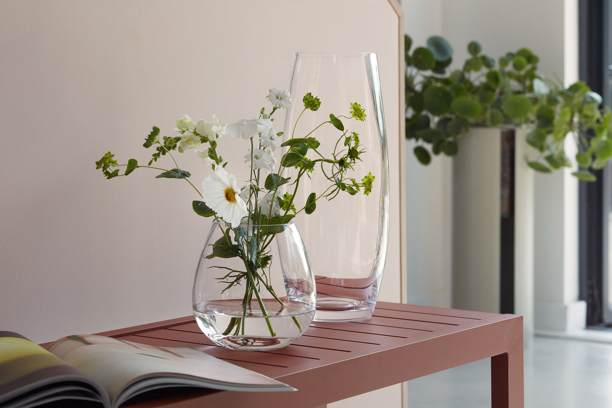 An lsa-international Barrel Bouquet Vase, featuring white flowers and greenery, sits atop a pink table beside a tall empty vase. In the foreground rests an open book, while a leafy plant is visible in the background.