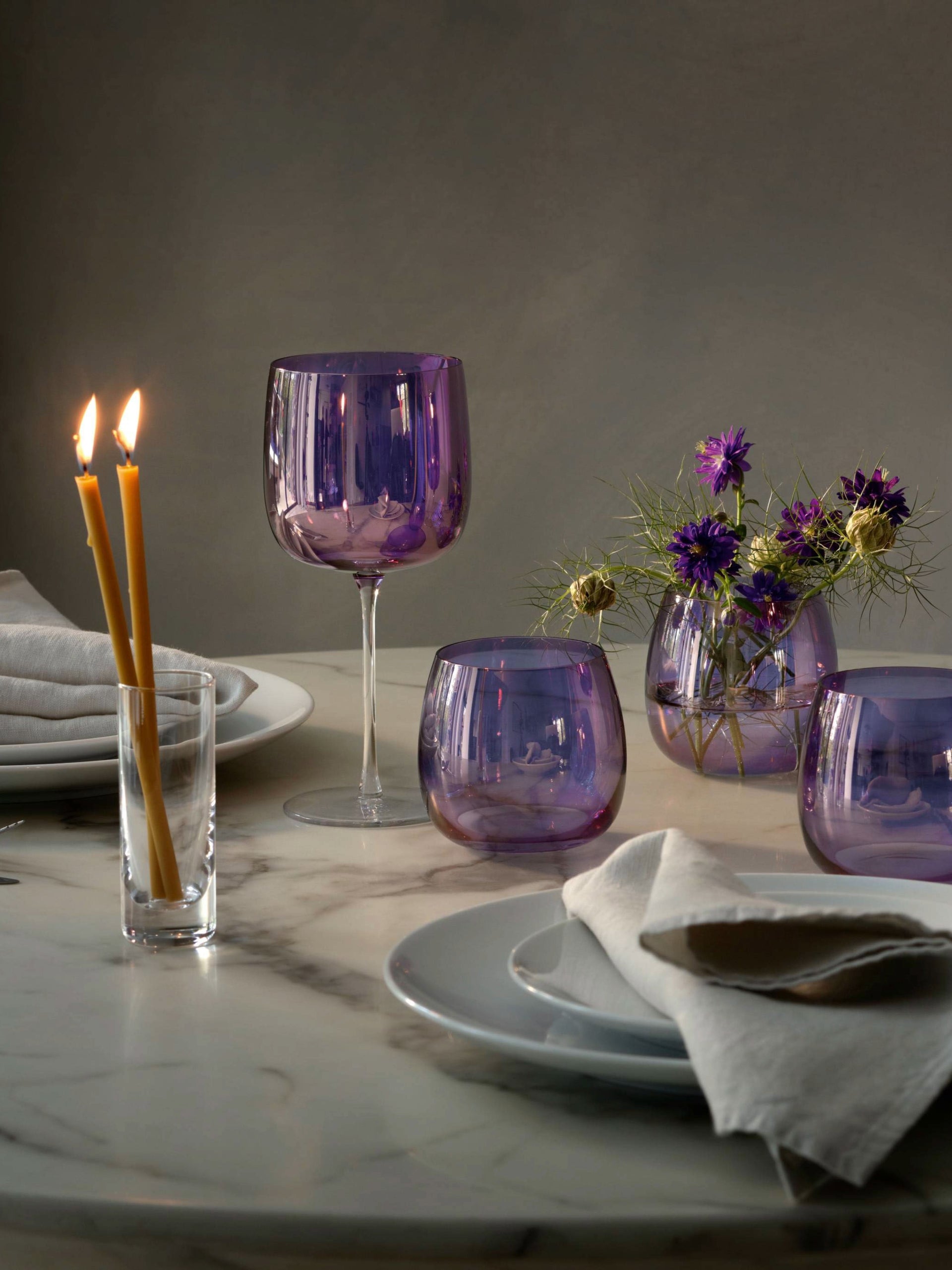 A marble table is elegantly set with plates, napkins, two lit taper candles in glass holders, and lsa-international Wine Glasses featuring a contemporary violet lustre, accompanied by a small vase of purple flowers. The scene is softly lit for a refined ambiance.