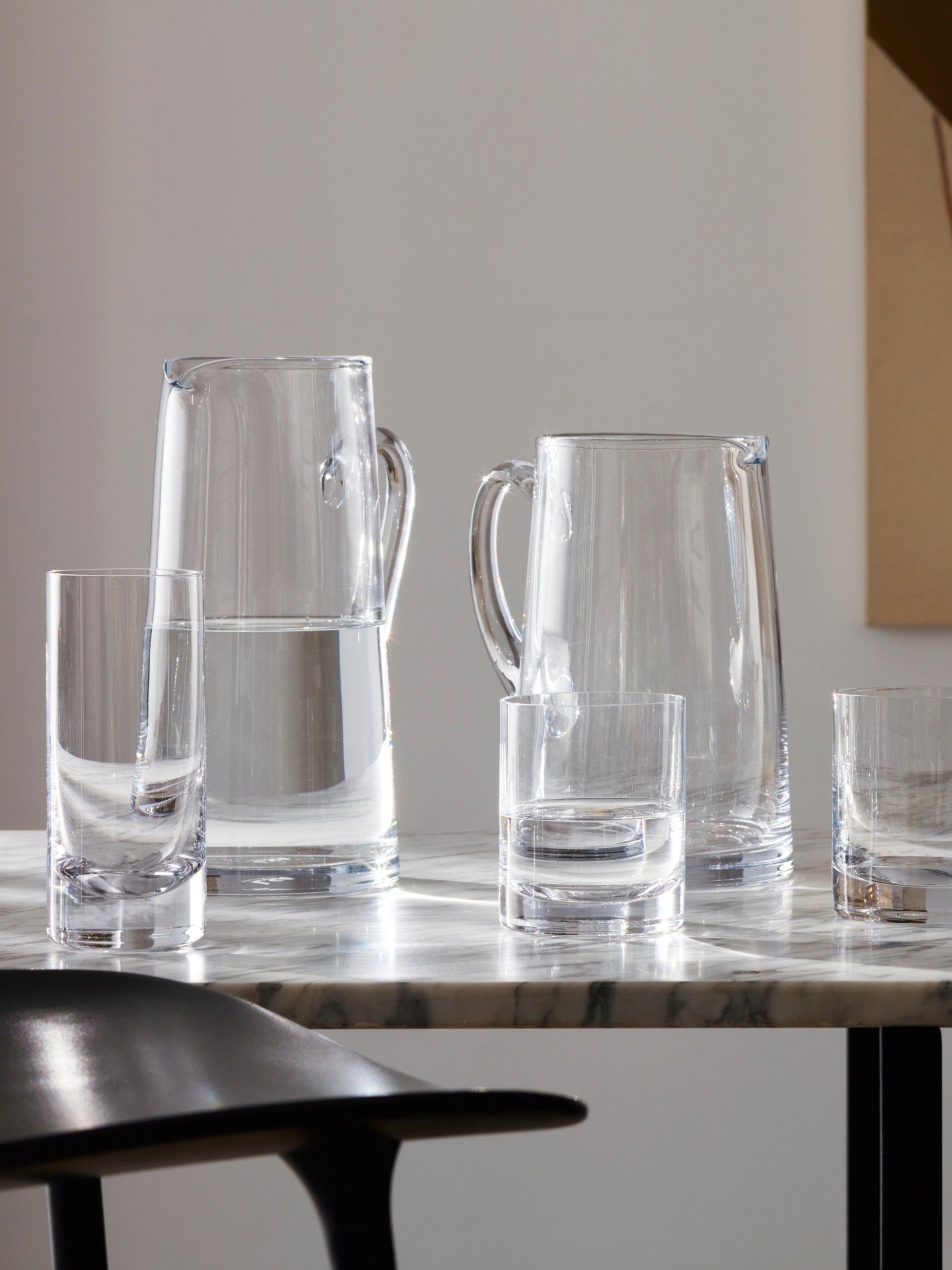 Two glass pitchers—each a mouth blown glass jug, partially filled with water—and three empty clear glasses are arranged on a marble table. Sunlight casts reflections and shadows, creating a clean, minimalist scene. A black chair is visible in the foreground.