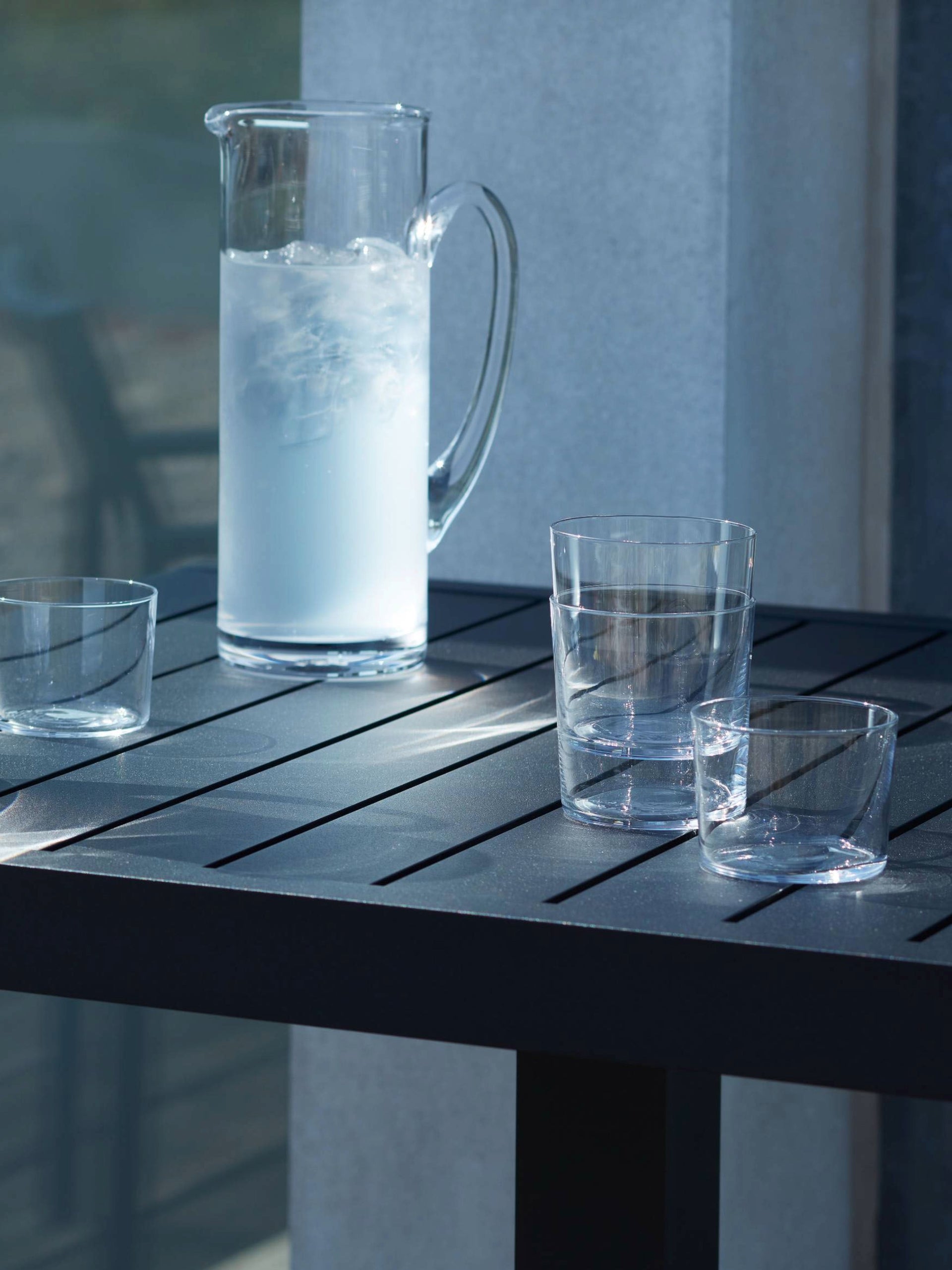 A mouth-blown lsa-international Jug filled with ice water and three empty glasses rest on a black slatted outdoor table. This elegant serving piece catches the sunlight, creating soft reflections on the chip-resistant finish of both the glassware and the table surface.