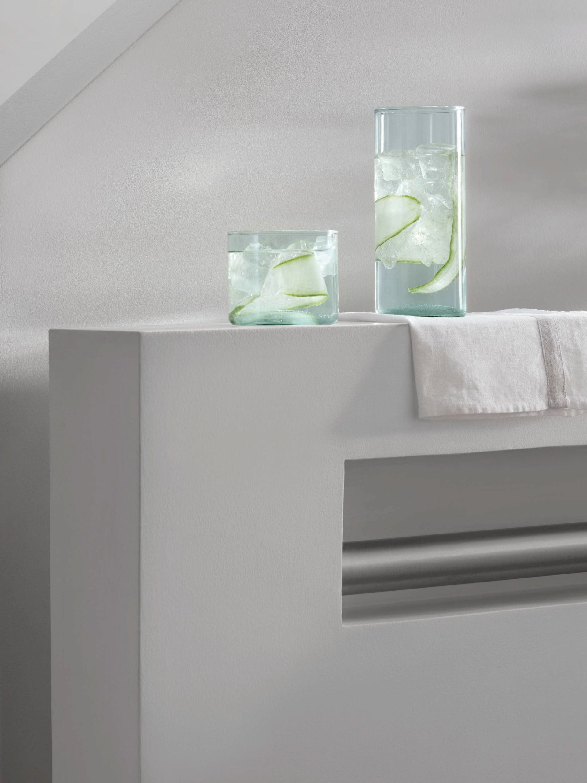 Two Highball glasses by lsa-international, crafted from clear, mouthblown recycled glass, are filled with ice, water, and cucumber slices. They rest on a white surface beside a folded white cloth in a minimal, modern setting—an ideal accent for the sustainable lifestyle.