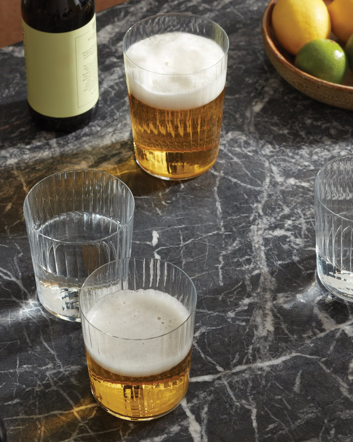 Two lsa-international Tumbler glasses filled with beer and two more holding water are set on a dark marble table beside a bottle and a bowl brimming with lemons and limes. These stackable Tumblers are both dishwasher safe, blending practicality with elegant design.