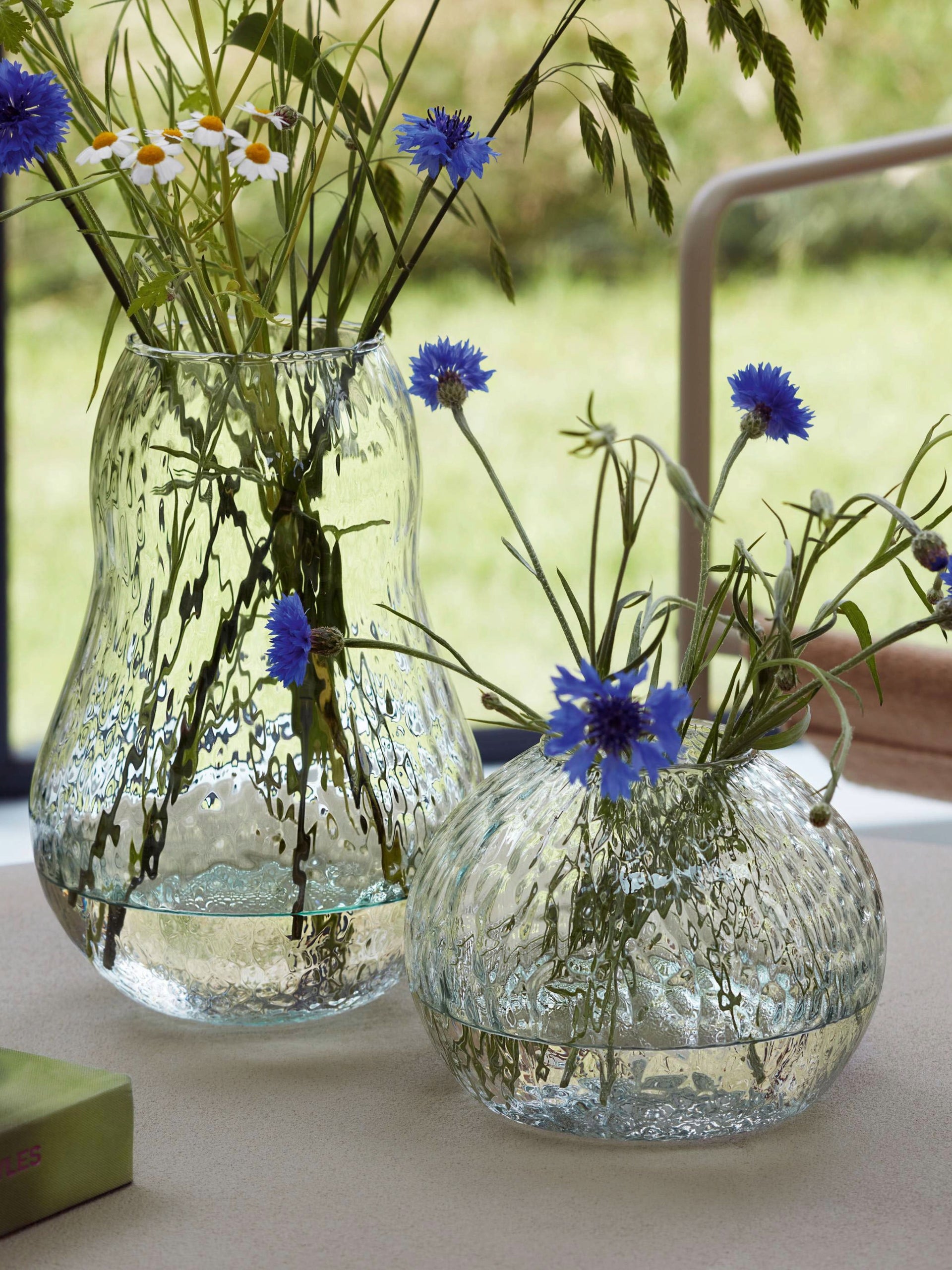 Two lsa-international Seed Vases made from textured recycled glass, each filled with wildflowers such as blue cornflowers and white daisies, are arranged on a light surface by a window, with a softly blurred green outdoor scene in the background.
