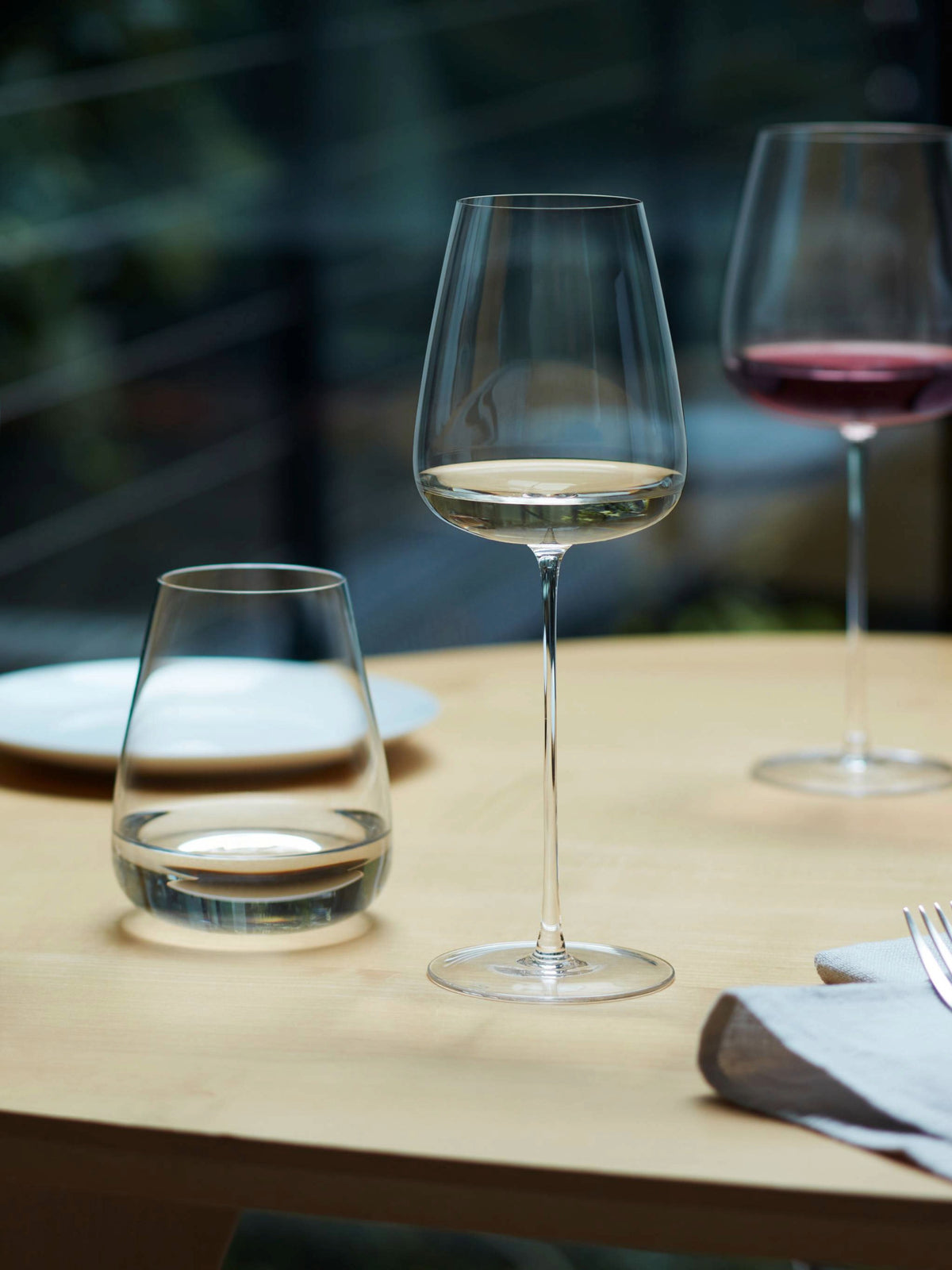 A table set with a hand-crafted, mouth-blown lsa-international White Wine Glass filled with white wine, a glass of water, a modern red wine glass in the background, a plate, a napkin, and a fork.