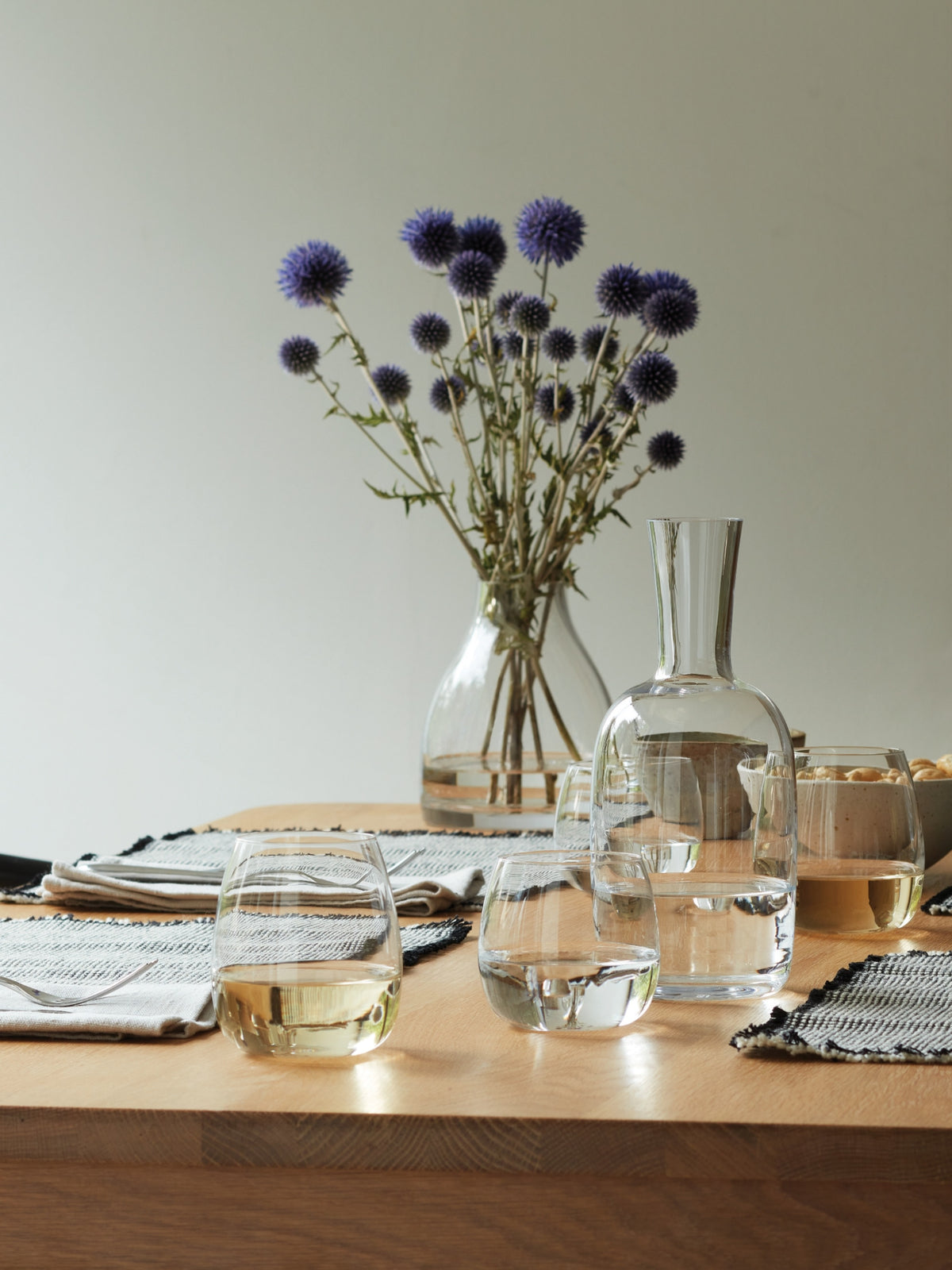 A wooden table set with lsa-international Stemless Glasses, a clear carafe, and placemats. In the background, a vase holds purple thistle flowers against a plain wall.