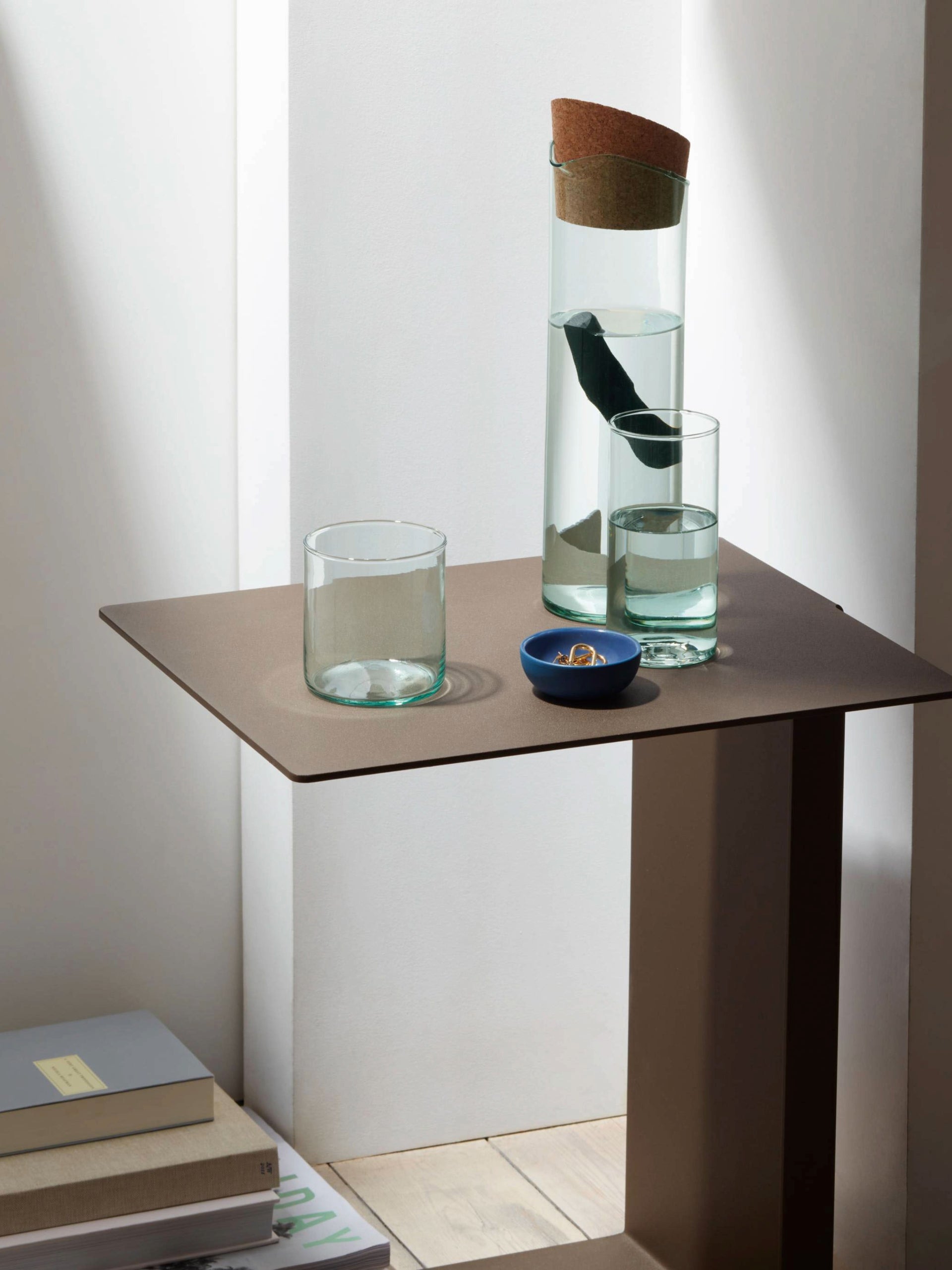 A minimalist table holds a mouthblown lsa-international Highball glass carafe with a cork, two lsa-international Highball glasses made from recycled glass filled with water, and a small blue dish holding rings. Soft, natural light casts gentle shadows on the sustainable scene.