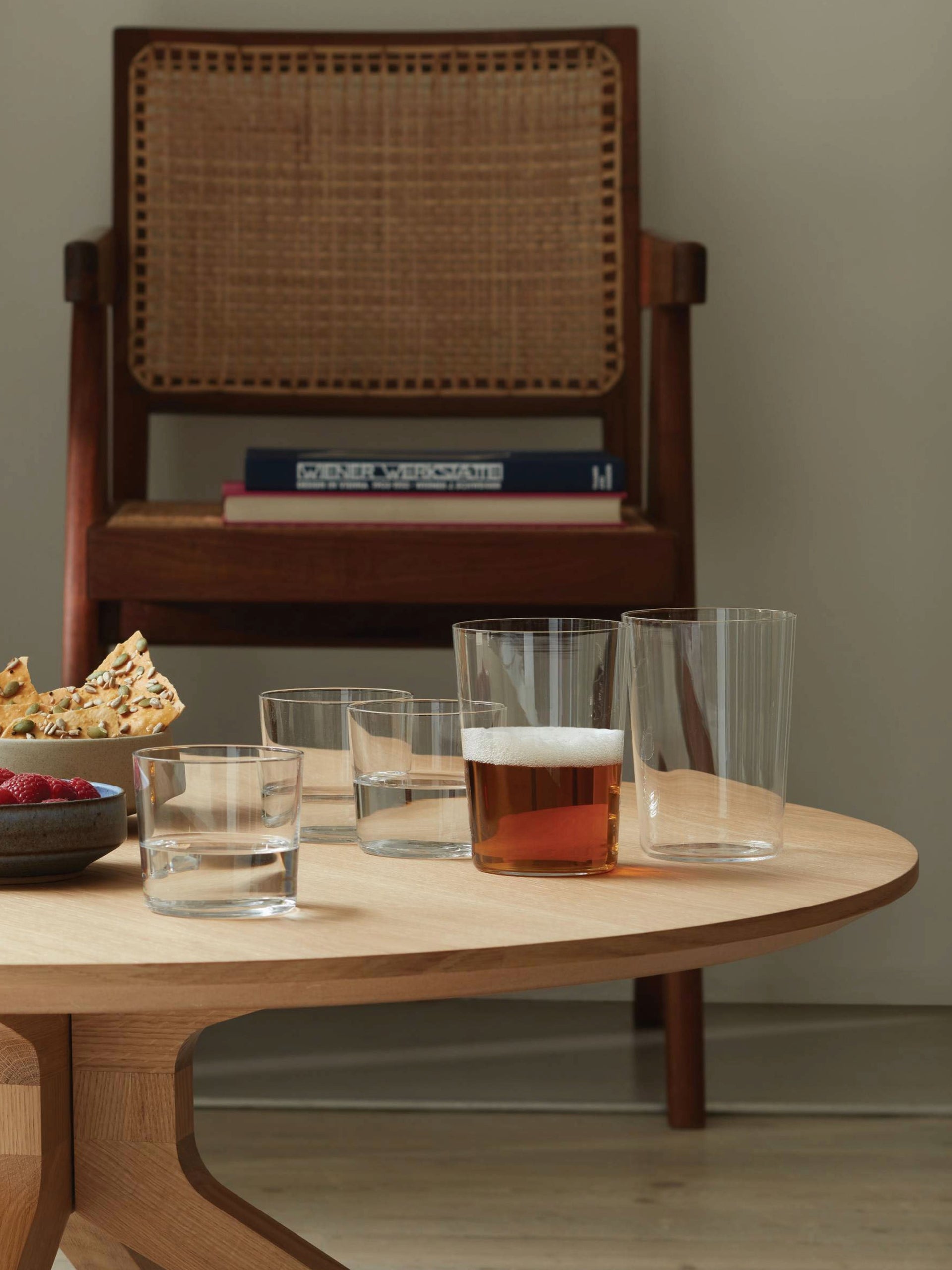 On a wooden table, there are stackable lsa-international Tumbler glassware filled with water, a glass of beer, a bowl of berries, and snacks, with books stacked on a woven-backrest chair in the background.