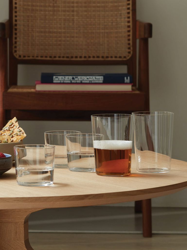 A set of clear glass drinking tumblers arranged on a light wood table in a real interior setting, with a wooden chair and books in the background.