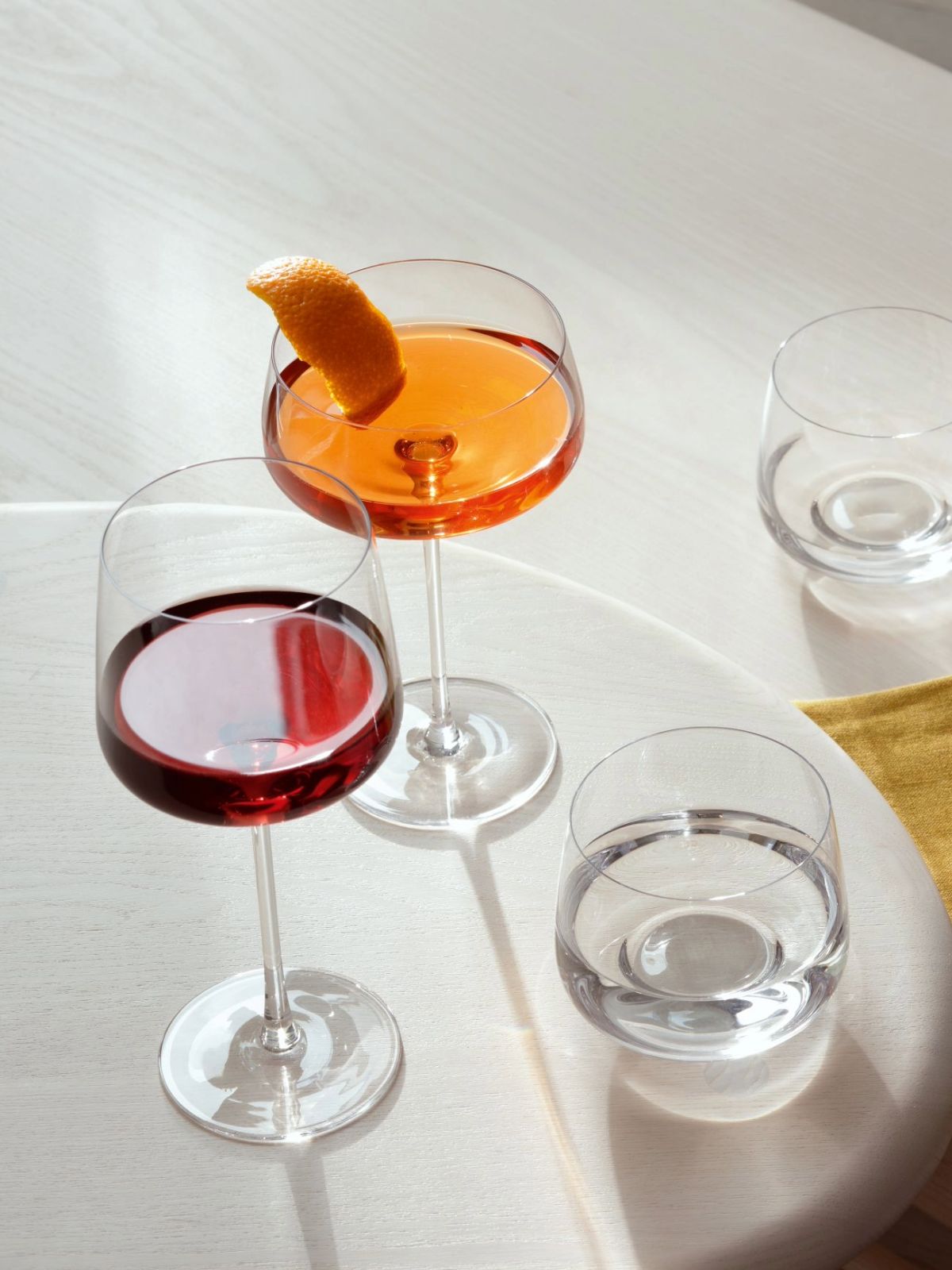 A styled tabletop arrangement of clear drinking glasses and stemmed wine glasses on a light surface, with orange and red liquids inside, a small glass bowl of nuts on a yellow napkin, and soft shadows across the scene.