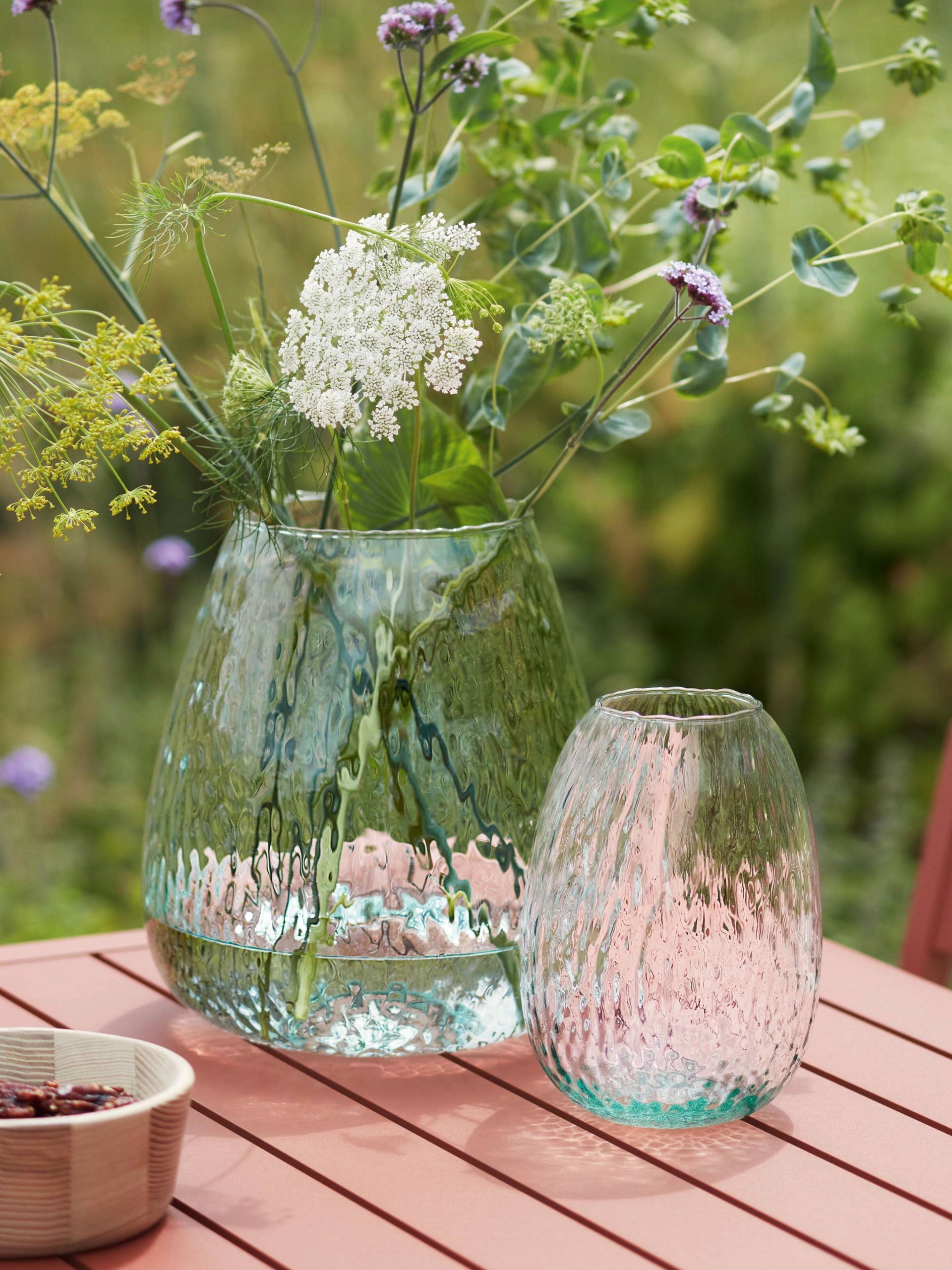 Two textured Seed Vases by lsa-international, one large and one small, sit on a pink outdoor table. The large vase holds assorted wildflowers in vibrant hues, against a blurred green garden backdrop.
