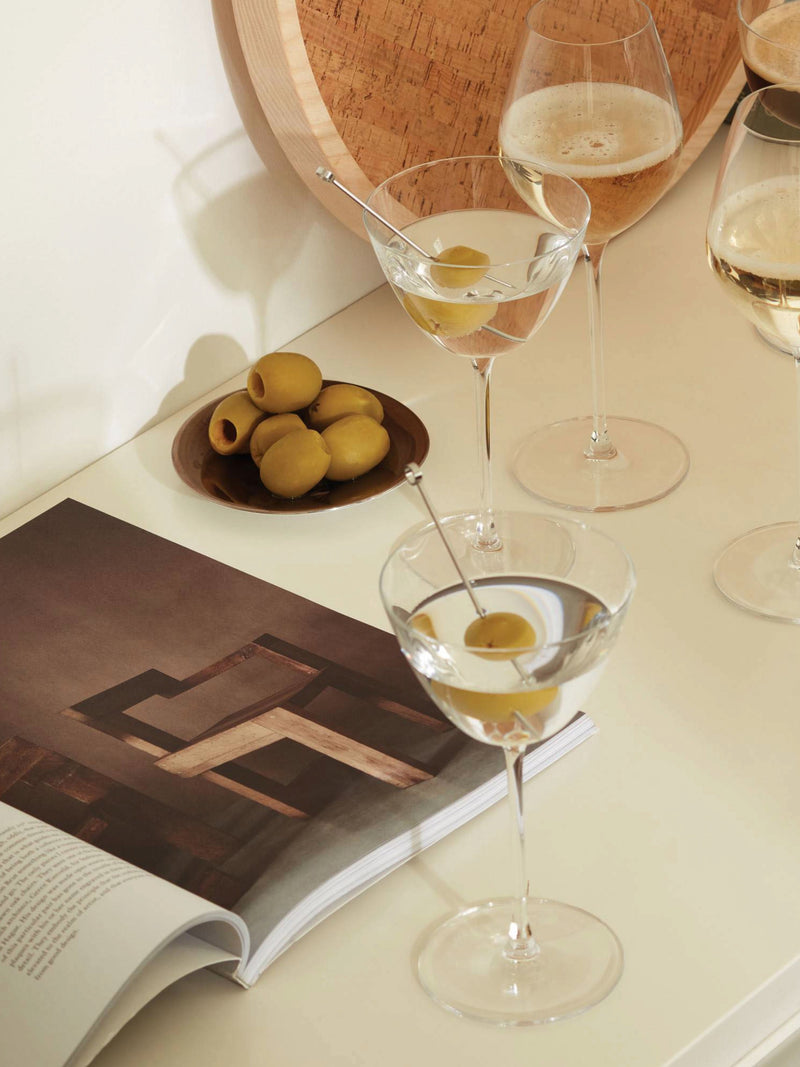 A tabletop scene showcases two lsa-international Martini Glasses—crafted from lead-free crystalline glass and garnished with green olives—next to a bowl of olives, three sparkling cocktail glasses filled with white wine or champagne, and an open book displaying a wooden chair. Warm, soft lighting bathes the arrangement in a cozy glow.