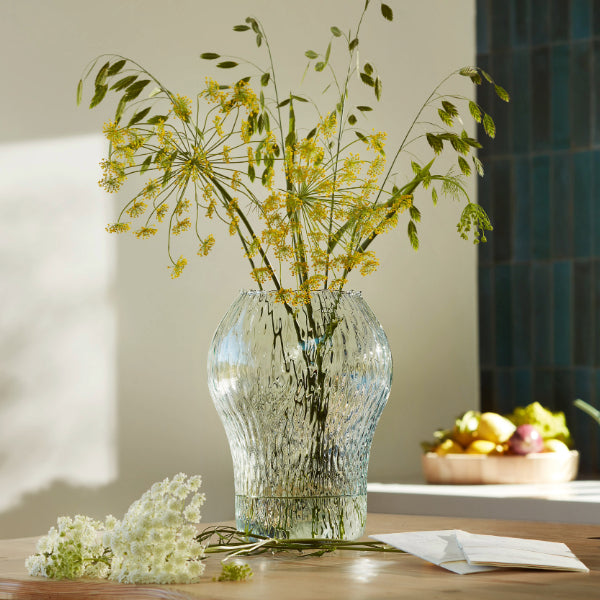A clear textured glass vase filled with yellow wildflowers and green stems, placed on a wooden table in a softly lit interior setting.