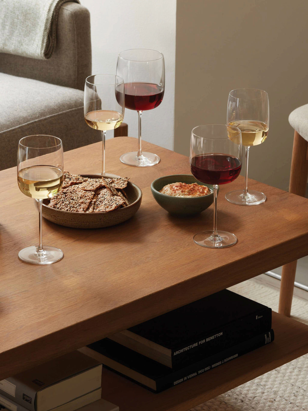 Four lsa-international Wine Glasses with tall stems, filled with red and white wine, are arranged alongside a plate of crackers and a bowl of dip on a wooden coffee table with books on the lower shelf, situated next to a couch and chair.
