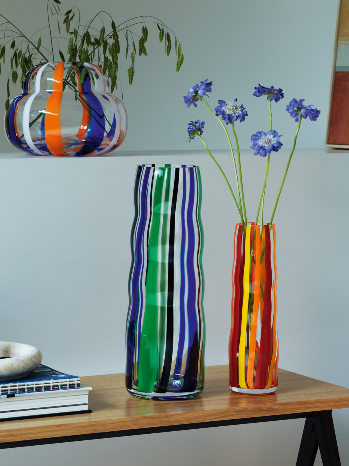 Three colorful, mouth-blown lsa-international Vase with wavy vertical stripes sit on a wooden table—one holds purple flowers, another is empty, and a rounder Vase in the back holds a leafy plant.