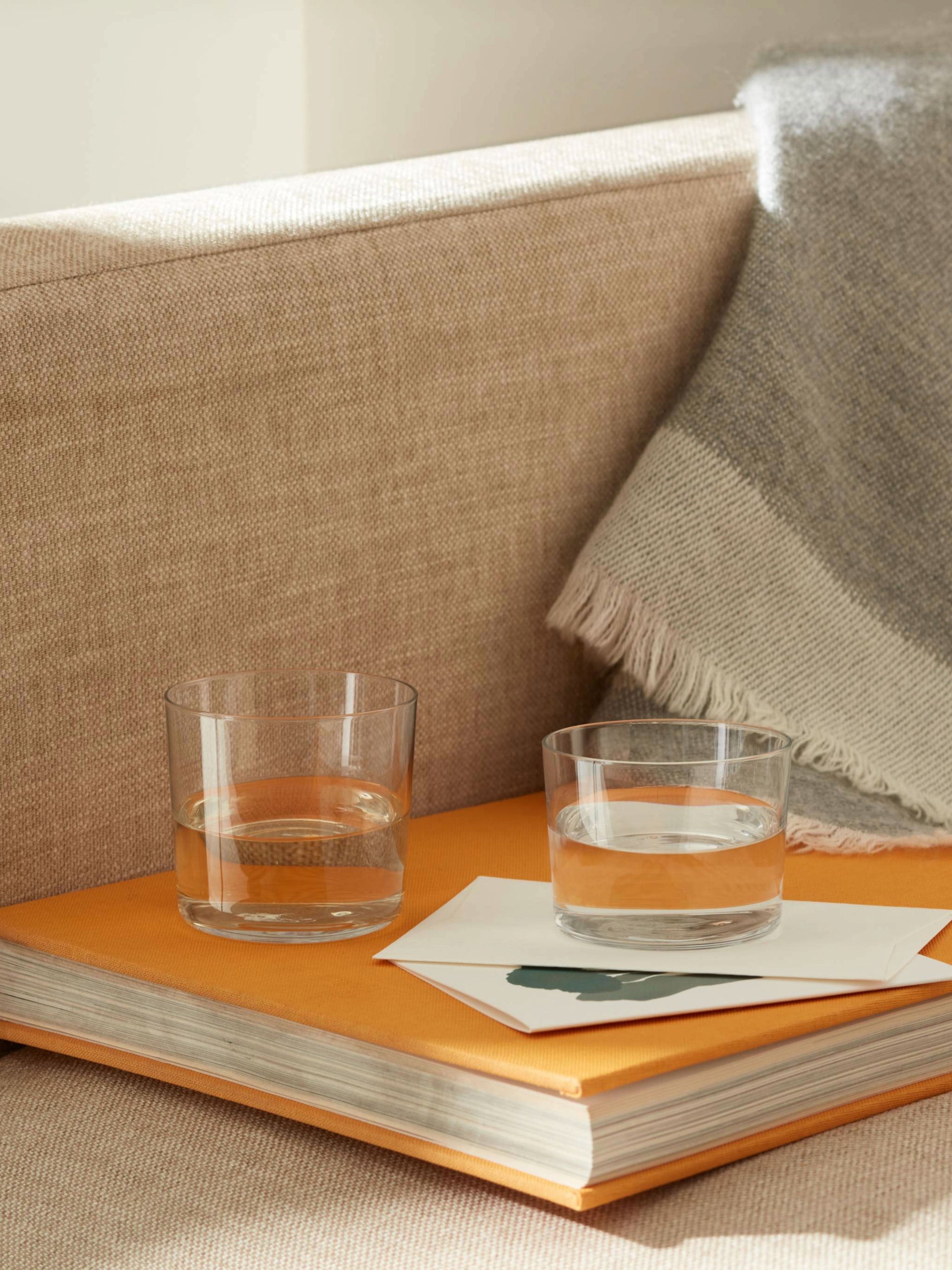 Two clear lsa-international Tumbler glasses filled with water rest atop a closed orange book beside a folded blanket draped over the edge of a beige couch, softly illuminated by sunlight.