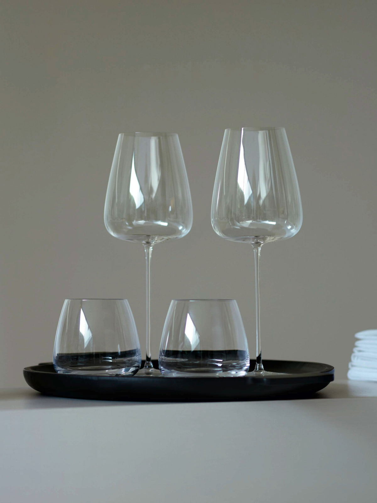Four empty glasses—two lsa-international Red Wine Grand Glasses and two short tumblers—are arranged on a round black tray. A small stack of white napkins is visible to the right. The background is a plain, neutral wall.