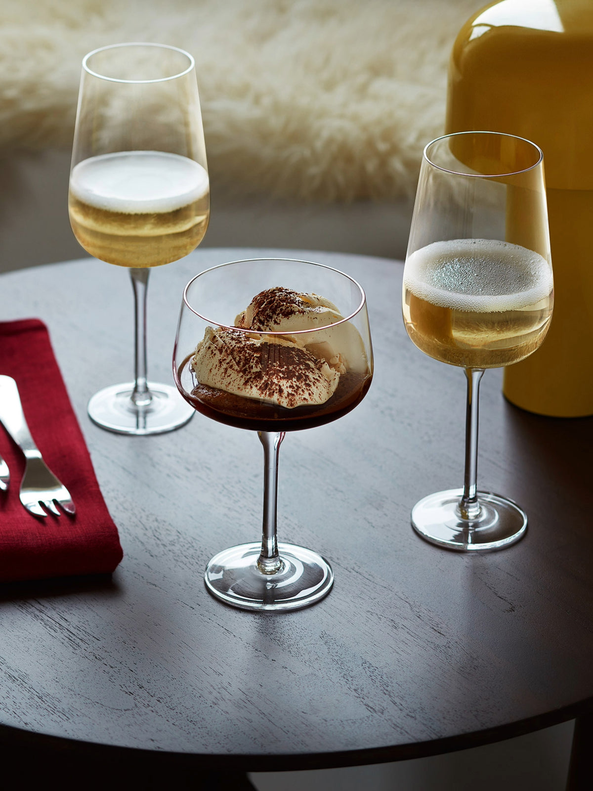 Three lsa-international Flute Glasses sit on a dark table: two filled with white wine and one elegantly presenting a dessert topped with cocoa powder. Ideal for daily use, this lead-free set is accented by a red napkin with cutlery and a yellow object nearby.