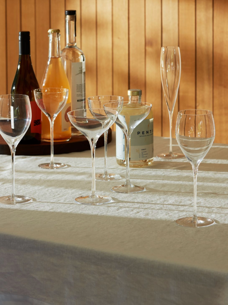 A sunlit table with a white cloth holds several empty wine and cocktail glasses. In the background, assorted bottles of alcohol are arranged against a wood-paneled wall.