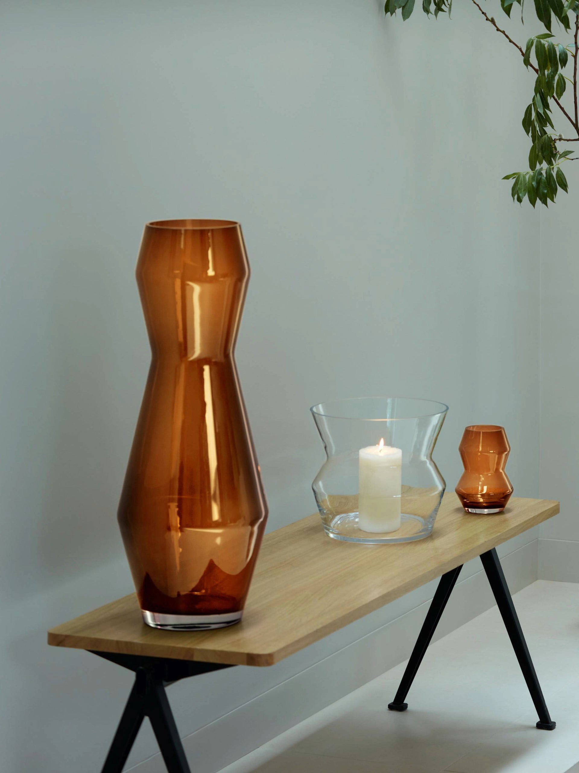 A wooden bench displays the lsa-international Vase, a modern mouth-blown design perfect for large bouquets, alongside a glass candle holder with a lit white candle and a small amber vase. A green plant hangs above on the right.