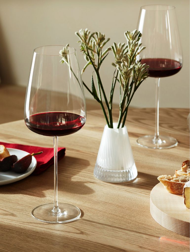 Two glasses of red wine on a wooden table, with a small vase of white flowers, plates with bread and cheese, and a red napkin in the background.