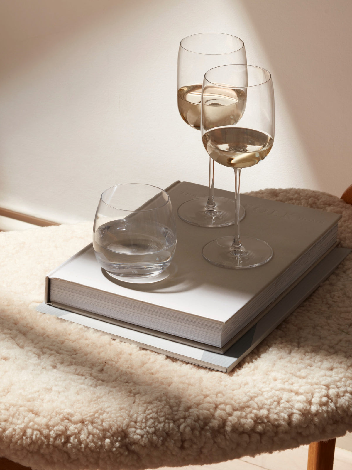 Two lsa-international Wine Glasses with tall stems, each filled with white wine, and a tumbler of water rest on a pair of stacked books placed atop a soft, cream-colored textured surface bathed in gentle natural light.