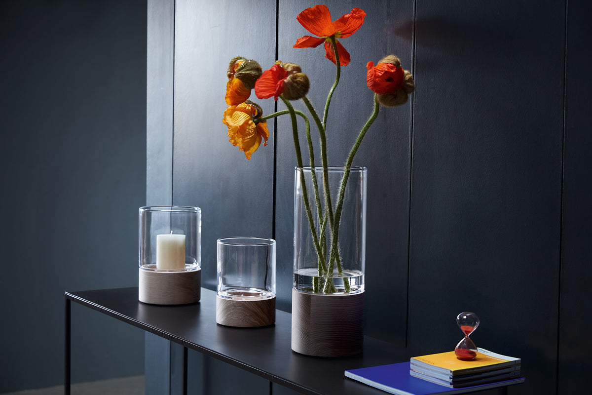 Three mouth-blown lsa-international Vases with elegant ash bases are arranged on a dark table; one holds an orange candle, one stands empty, and one displays vibrant orange flowers. Stacked books and a small red hourglass sit nearby, all set before a dark, textured wall.