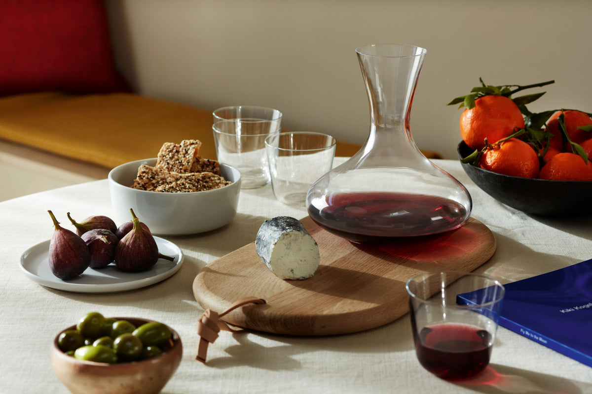 A white tablecloth displays the lsa-international Carafe & Oak Cheese Board arranged with a glass carafe filled with red wine, figs, cheeses on the oak board, olives, crackers, a bowl of mandarins, drinking glasses, and a blue book.