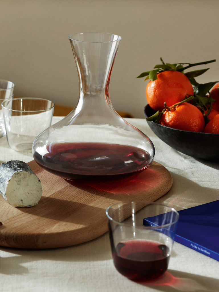 A clear glass decanter filled with red wine sits on a round wooden board on a table, with a glass, a blue book, a sliced cheese, and a bowl of oranges in the background.