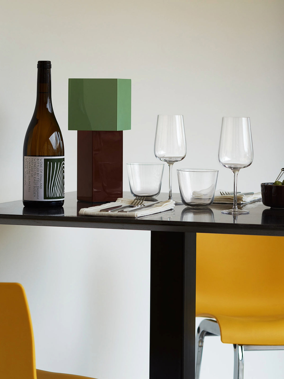 A modern dining table set with a wine bottle, a geometric green and brown accent piece, lsa-international Flute Glasses made from lead-free glass, utensils, napkins, and yellow chairs against a plain background—perfect for daily use.