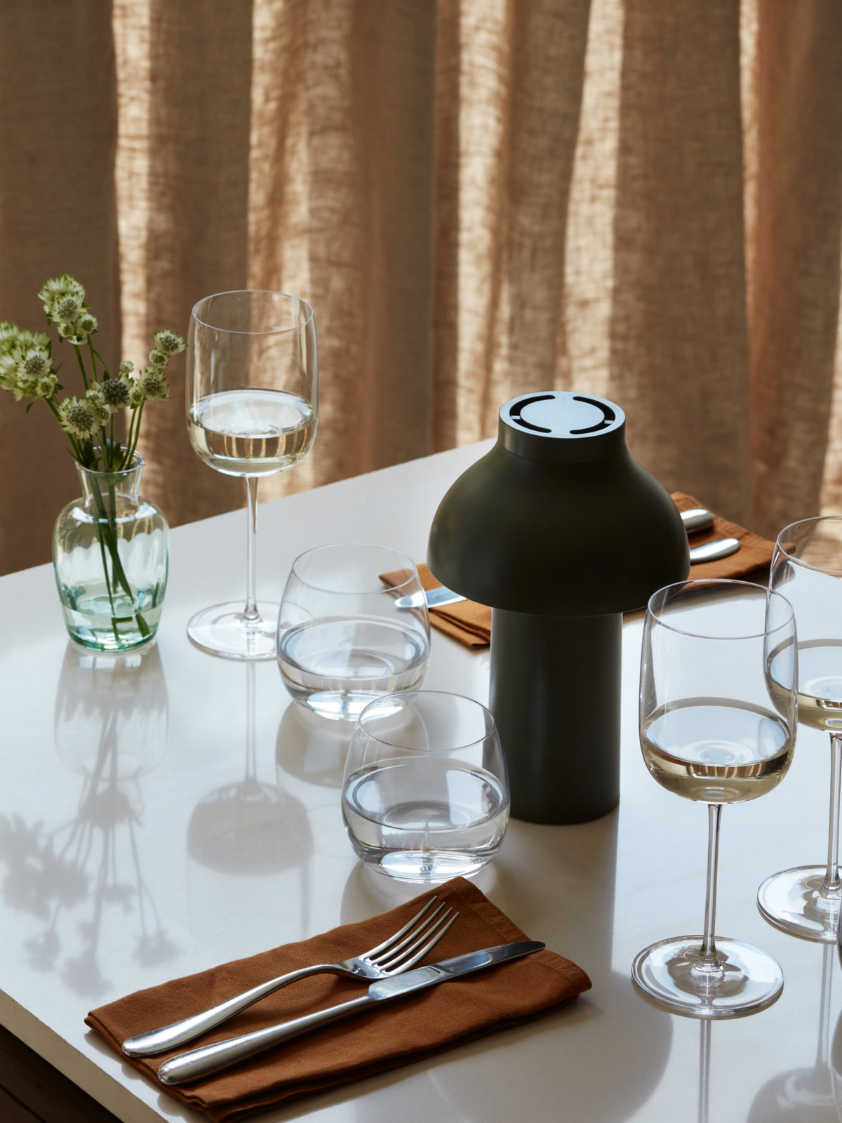 A set dining table featuring lsa-international Wine Glasses with tall stems, accompanied by water glasses, orange napkins with silverware, a green vase filled with white flowers, and a modern green lamp—all arranged on a white surface against beige curtains.
