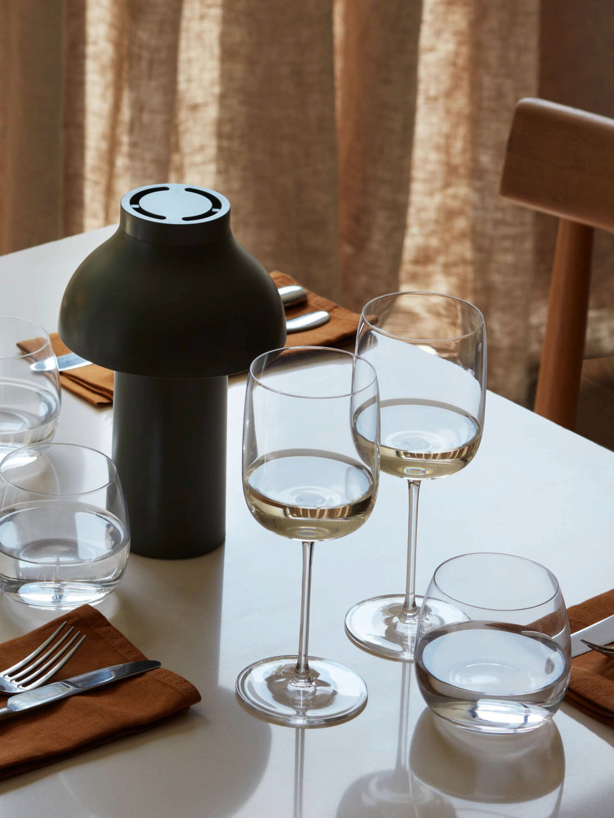 A set dining table with three lsa-international Wine Glasses featuring elegant tall stems, two water glasses, a dark modern lamp, brown napkins with cutlery, and neutral curtains in the background.