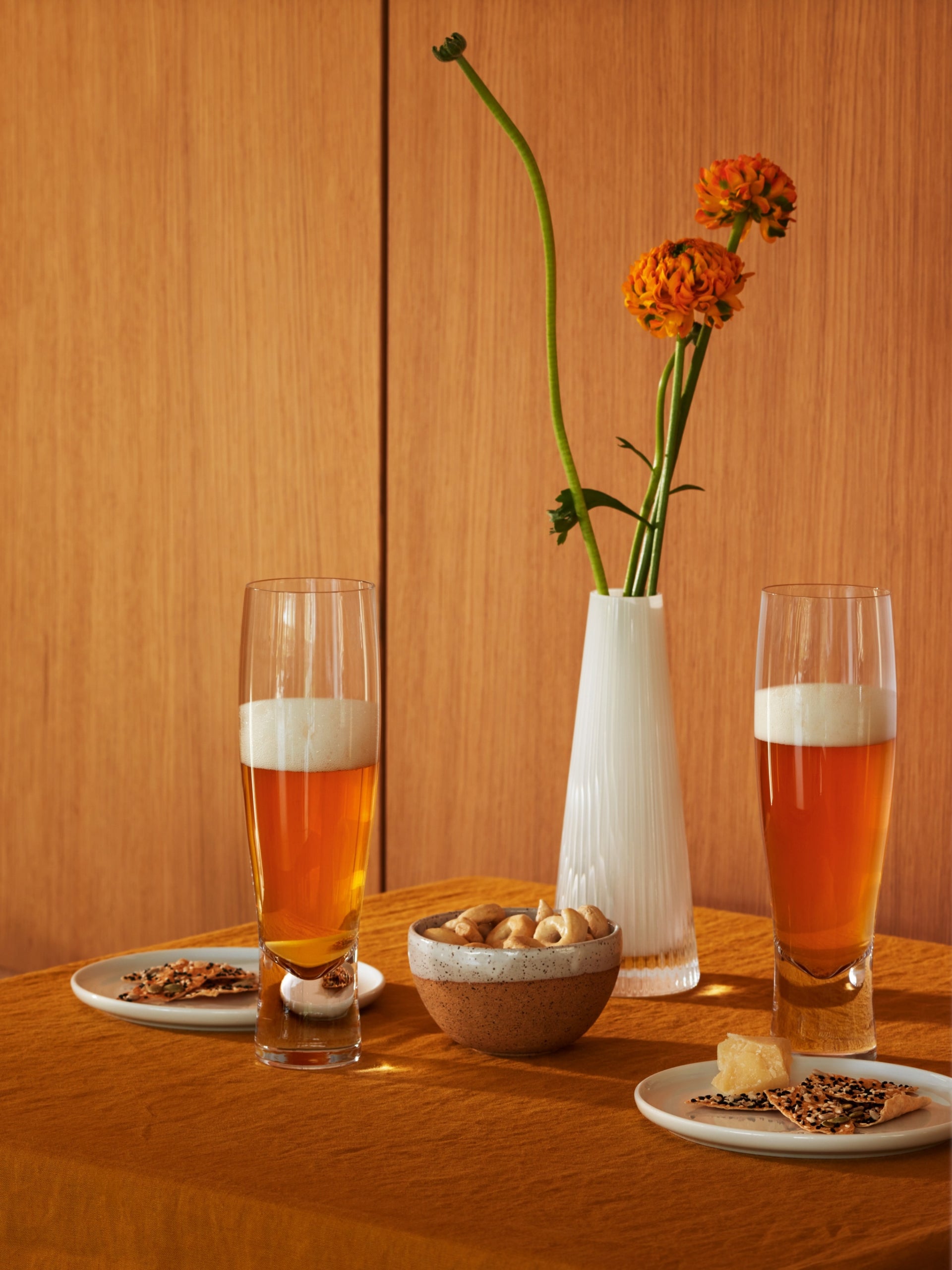Two lsa-international Lager Glasses, expertly crafted using the mouth-blown technique by skilled artisans, are filled with amber beer and set on an orange tablecloth beside a bowl of mixed nuts and two plates with assorted snacks. In the background, a white vase with two orange flowers stands out against a wooden wall.