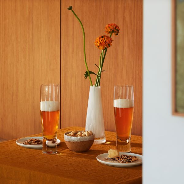 Two lager glasses filled with refreshing foamy beer, arranged on a wooden table with small plates, a bowl of nuts, and a white vase holding orange flowers against a wood-paneled wall.