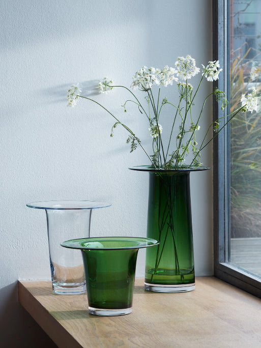 Three green and clear vases arranged on a wooden table next to the window with white wildflowers in the tallest vase, against a light gray wall.