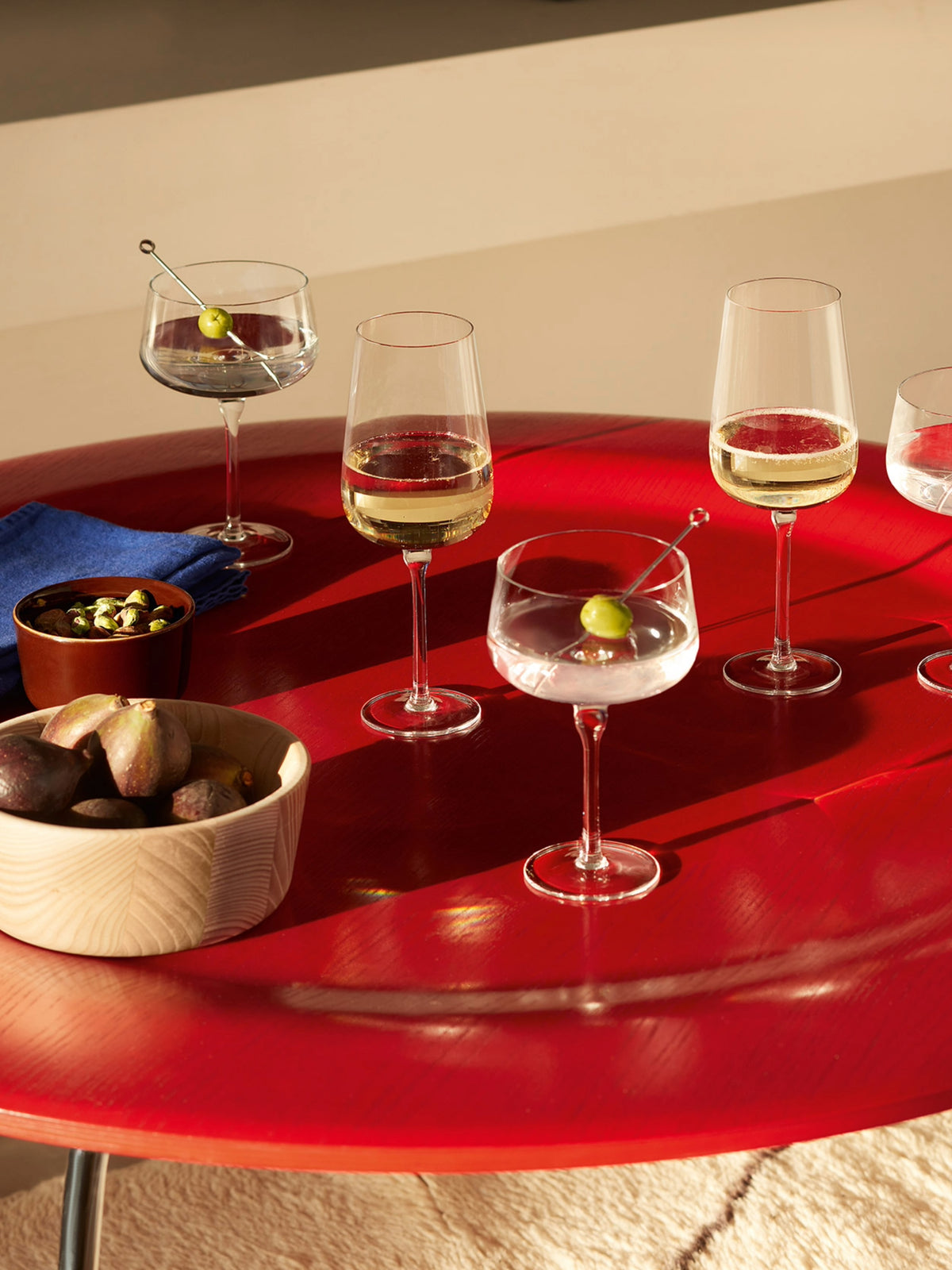 Five glasses of white wine and cocktails are elegantly served in lsa-international Flute Glasses, all arranged on a shiny red table alongside a blue napkin, a small bowl of nuts, and a wooden bowl of figs. Sunlight casts inviting shadows across the scene, creating an ideal setting for both daily use or gatherings.