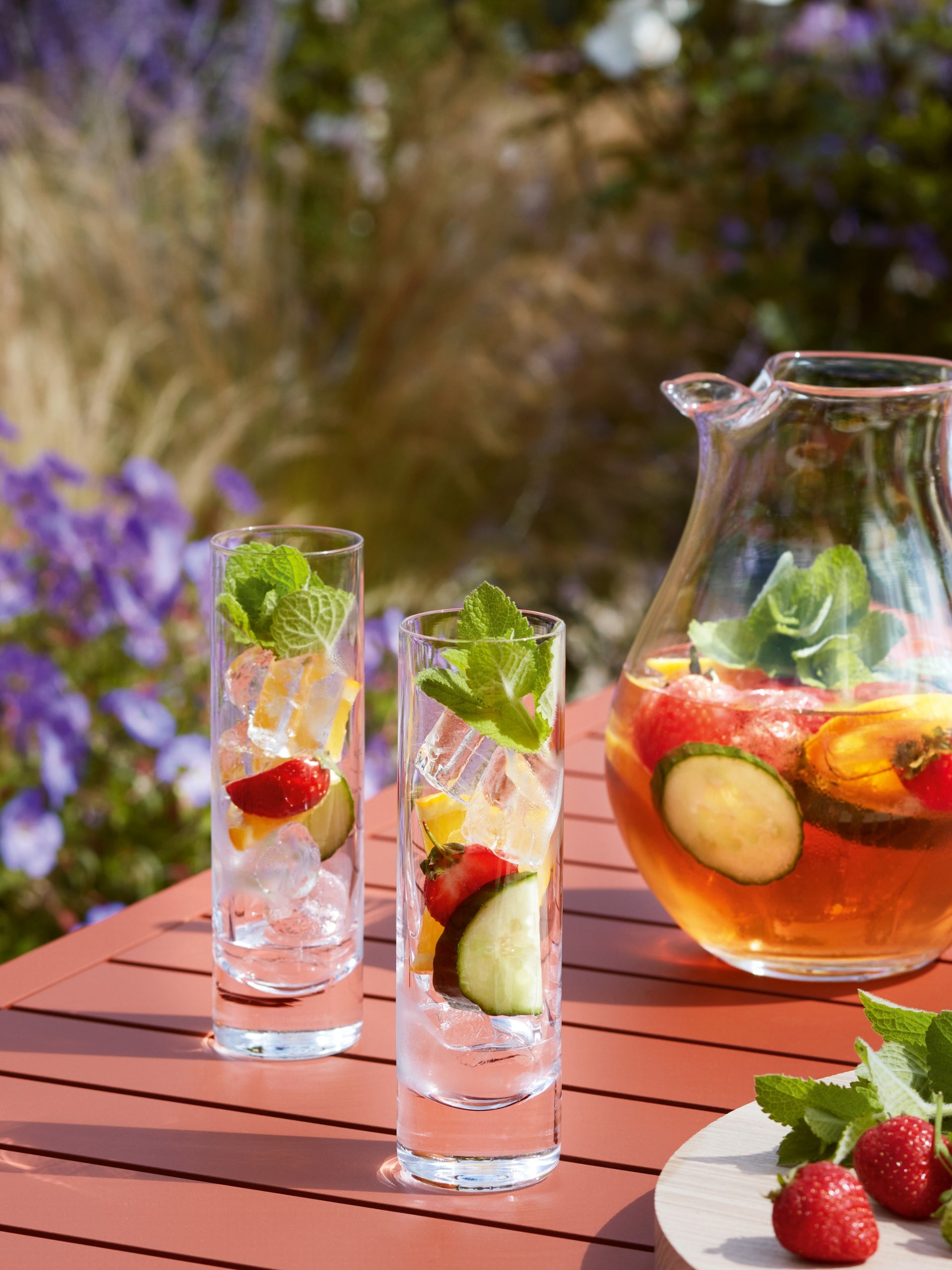 Two tall lsa-international Long Drink Glasses and a pitcher of fruity iced drink with cucumber, lemon, and strawberries sit on a red outdoor table; each Long Drink Glass is crafted from elegant mouth blown glassware and garnished with mint leaves, set against a blurred garden background.