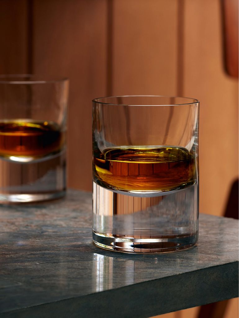 Two clear glasses filled with brown liquor sit on a gray stone surface, with one glass in focus and the other blurred in the background against a wooden wall.