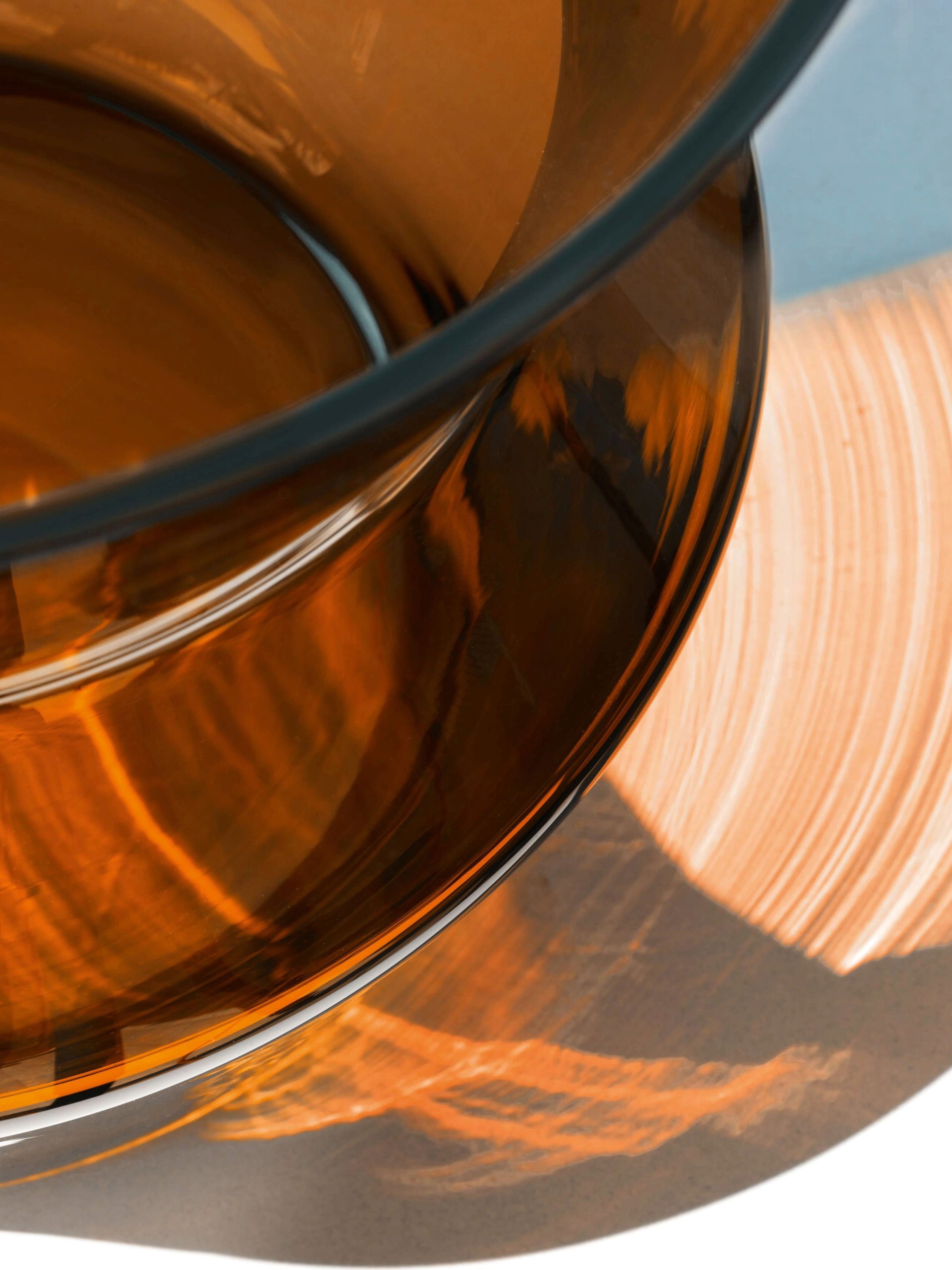 Close-up of a modern, mouth-blown Vase by lsa-international in amber glass, casting warm, abstract shadows and reflections on a white surface with light blue accents. Ideal as a tall vase or for large bouquets, its unique form adds contemporary elegance to any setting.