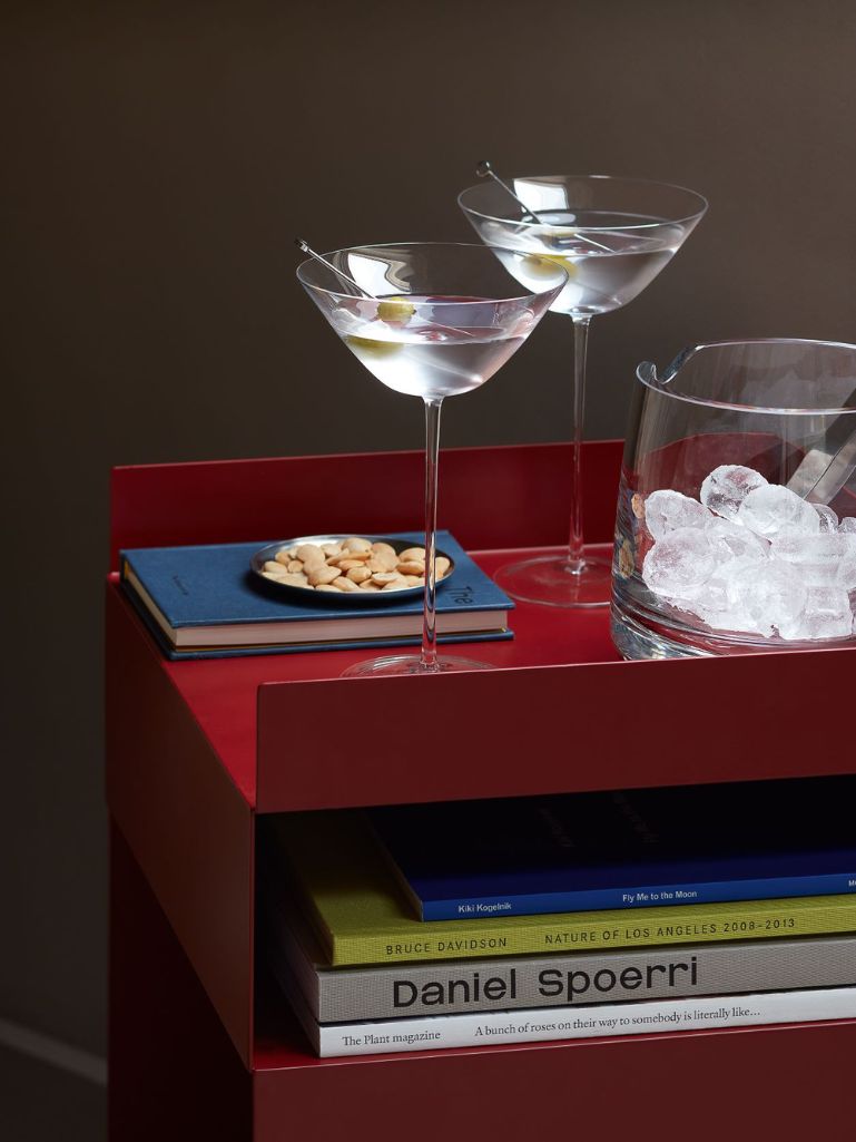 A red side table styled with two martini glasses, a glass bowl of ice, a small plate of nuts, and books, including a Daniel Spoerri book.