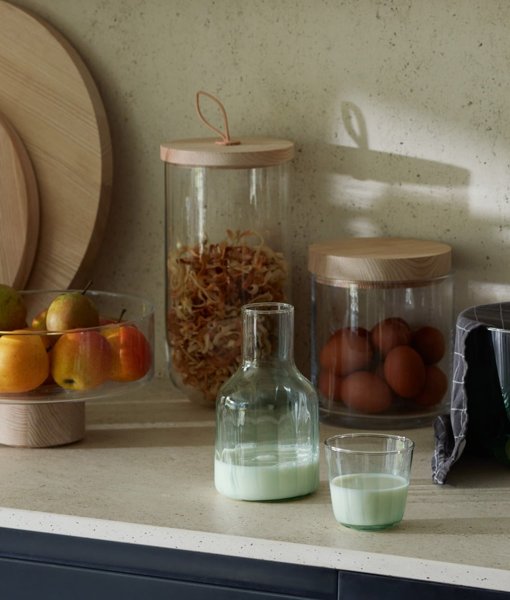A countertop holds a carafe and glass with milk, a bowl of apples, jars with eggs and dried food, and wooden boards, all in natural light.