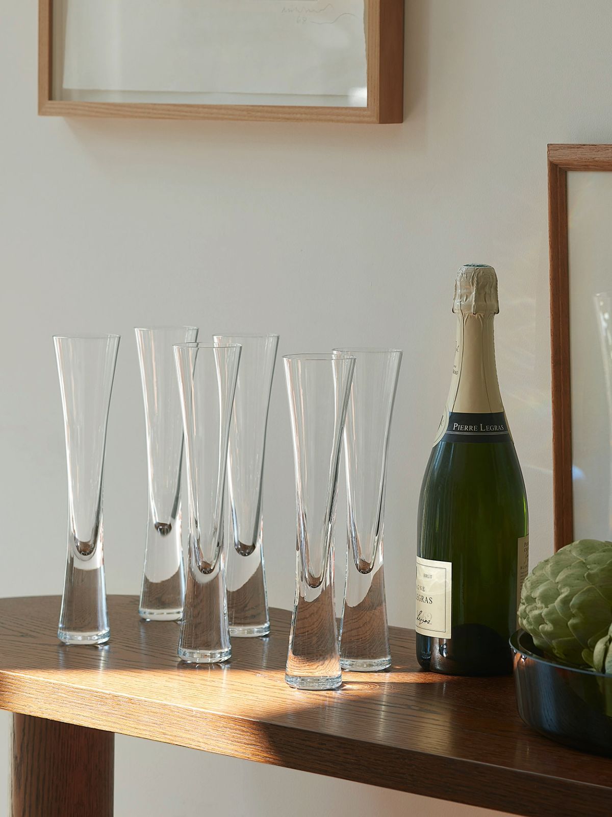 Six clear champagne flutes arranged on a wooden table beside a green champagne bottle and a bowl of citrus fruits, with a softly blurred indoor background.