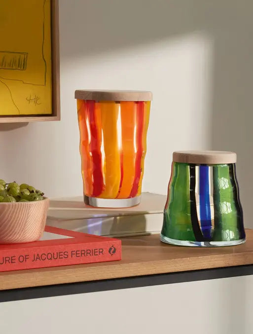 Two colorful glass jars with wooden lids sit on a shelf next to a bowl of green grapes and a red book titled JACQUES FERRIER. One jar is orange and yellow, the other is green with blue and black stripes.