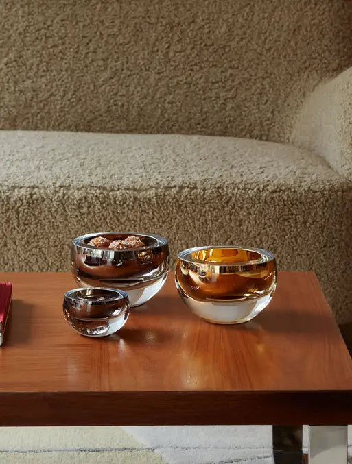 Three glass bowls with silver and gold metallic rims sit on a wooden table. One bowl contains wrapped chocolates. A fuzzy beige couch is in the background.