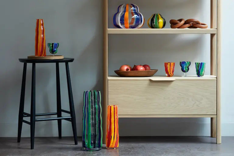 Colorful striped glass vases and cups are displayed on and beside light wooden shelves and a black stool. A wooden bowl with red apples and a wooden ornament are also on the shelf. The setting appears modern and stylish.