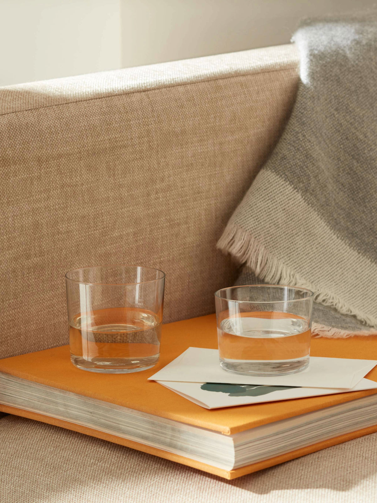 Two clear lsa-international Tumbler glasses filled with water rest atop a closed orange book beside a folded blanket draped over the edge of a beige couch, softly illuminated by sunlight.