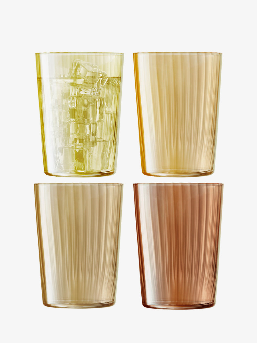 Four lsa-international Tumbler glasses, featuring ribbed fluted designs in yellow and amber hues, are arranged in a two-by-two grid on a white background; the top left glass contains ice cubes, while the others remain empty.